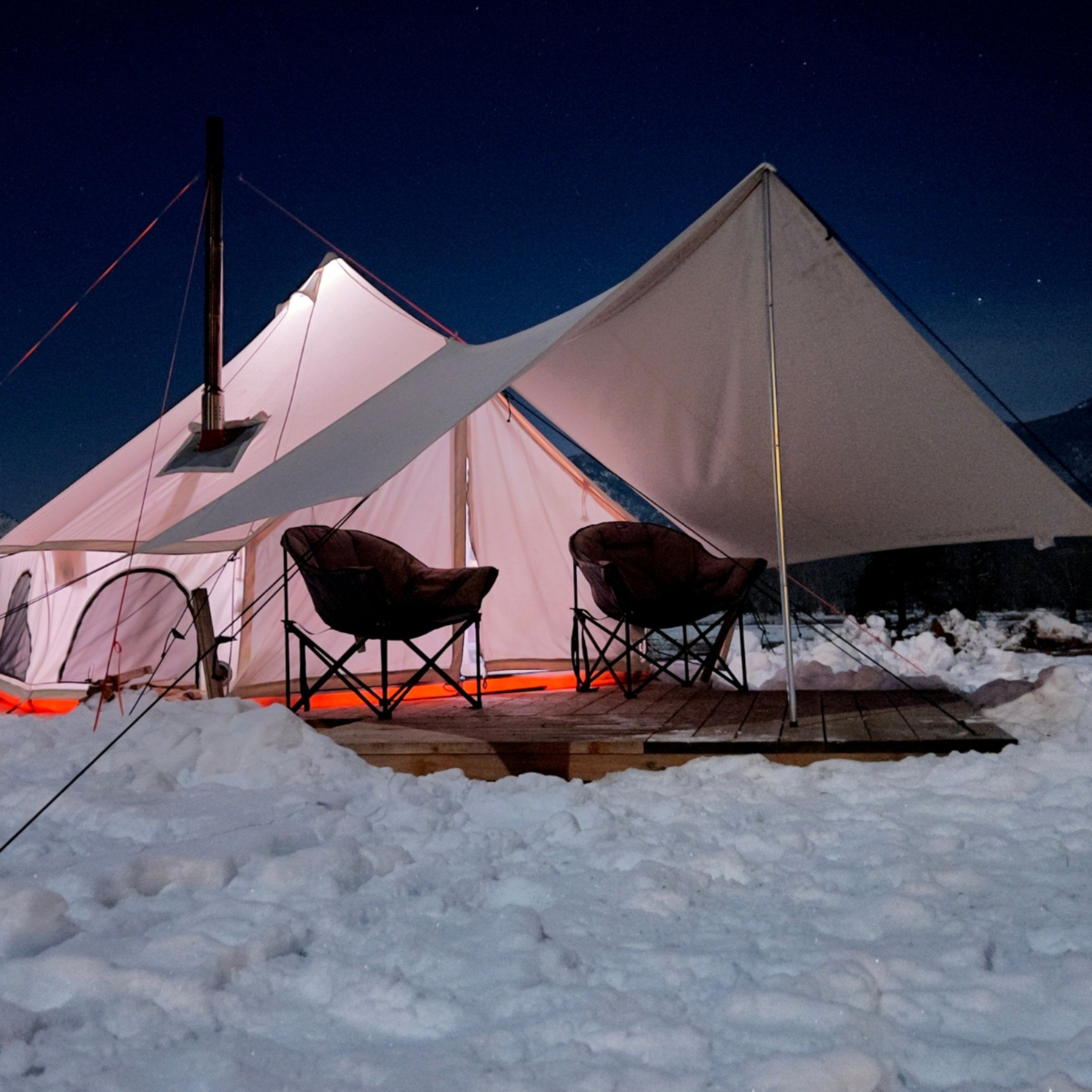 avalon bell tent set up in the snow on a platform with awning in front and camping chairs under it