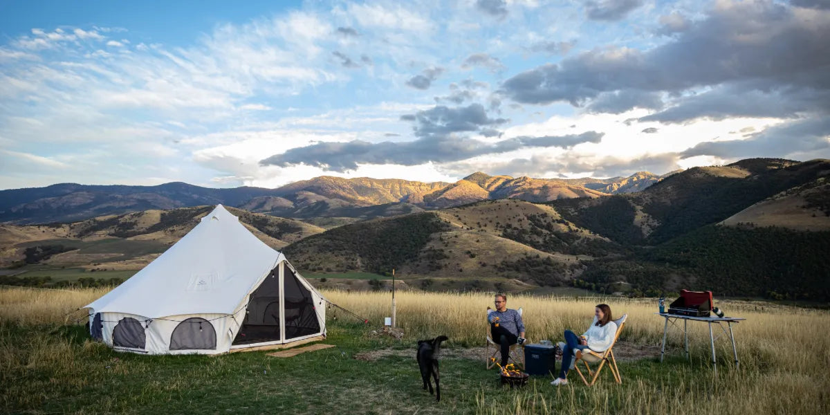 Couple relaxing beside a white canvas bell tent with a campfire and dog in a scenic mountain valley during sunset camping adventure