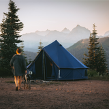 10' Regatta Bell Tent - Sapphire Blue placed in a forest with a woman and her dog walking towards it