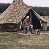 Regatta Bell Tent - Realtree Edge set up outdoors with a man sitting on a log right in front of it