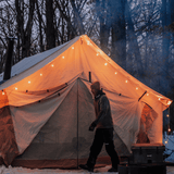 alpha wall tent in the snow with fairy lights glowing in the dark and a man walking in front of it